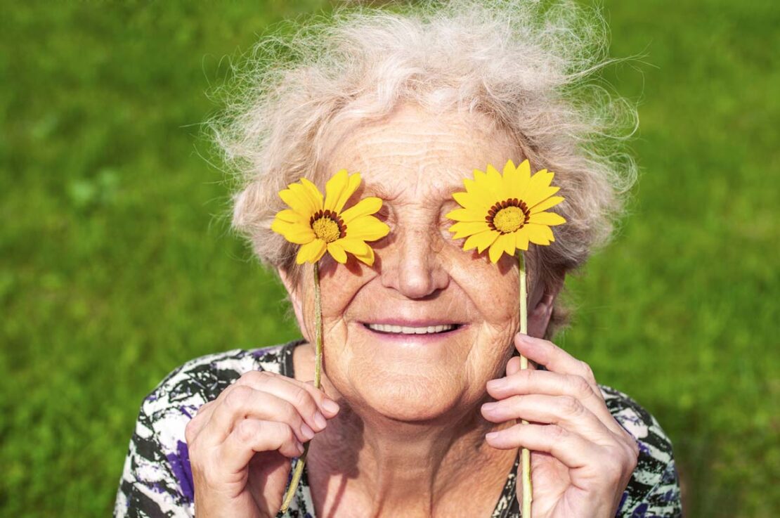 Una donna anziana sorride, tenendo due fiori gialli davanti agli occhi.