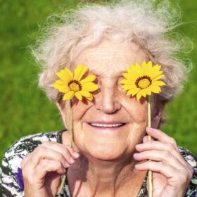 Una donna anziana sorride, tenendo due fiori gialli davanti agli occhi.